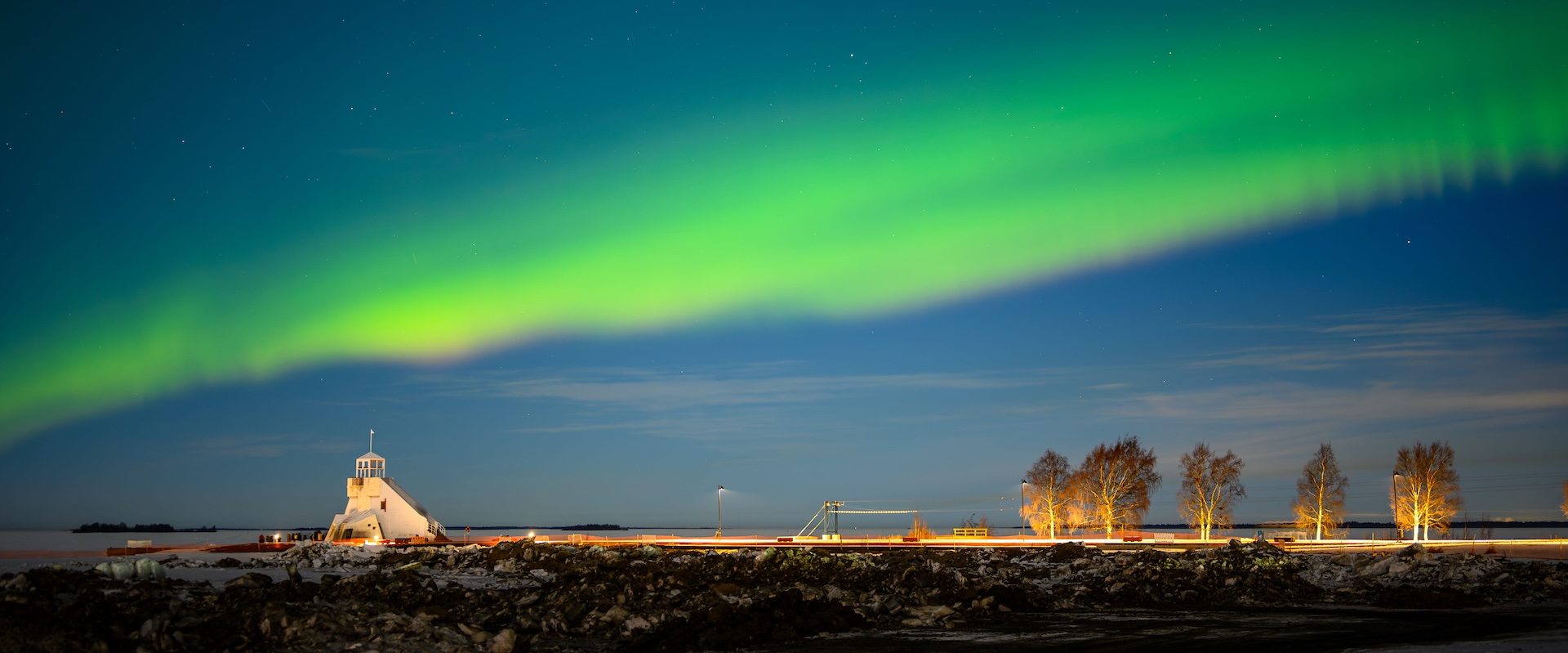 Green northern lights sweeping across the night sky over the frozen Oulu seafront, a lit waterfront tower and bare birches below. Photo: Peter Kurzok
