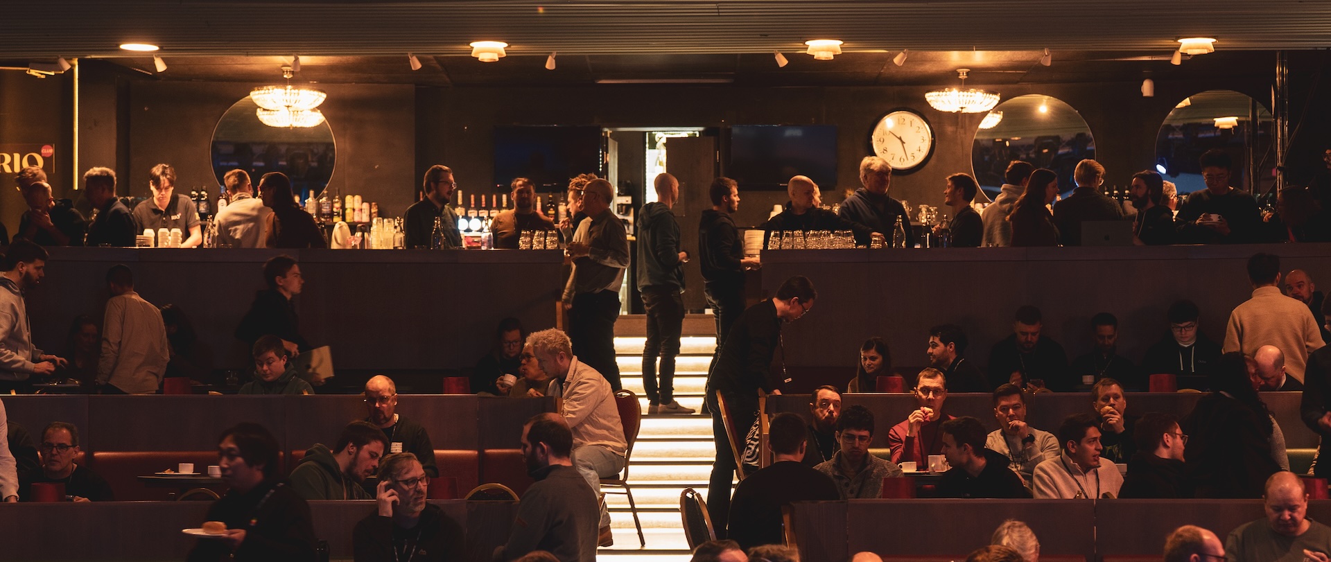 Conference-goers mingling over drinks during a break in the tiered Rio hall, a lit bar and clock along the back wall. Photo: Vera Lakovaara