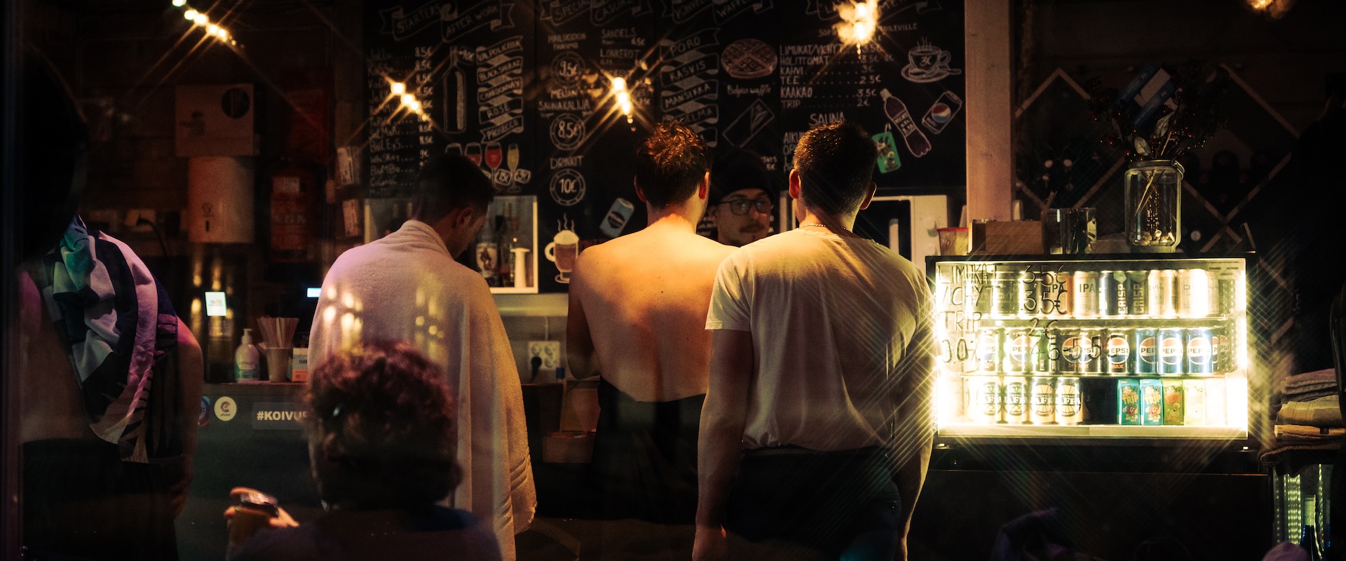 People in a warmly lit riverside sauna bar, one fresh from the sauna in a towel, ordering at a chalkboard-menu counter. Photo: Vera Lakovaara