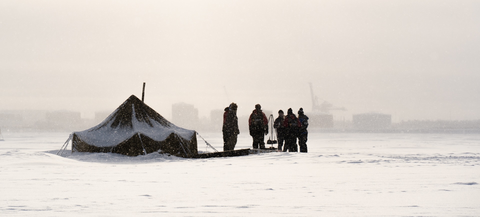A snow-covered tent on the frozen sea, a small group in winter gear beside it as snow falls, Oulu's harbour hazy in the distance. Photo: Vera Lakovaara
